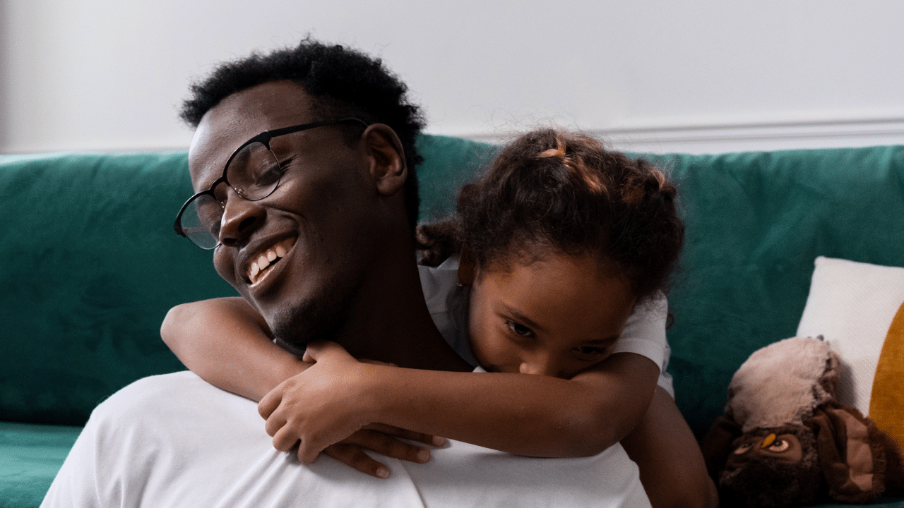 Young child with braids sits on a green couch and hugs her dads neck.