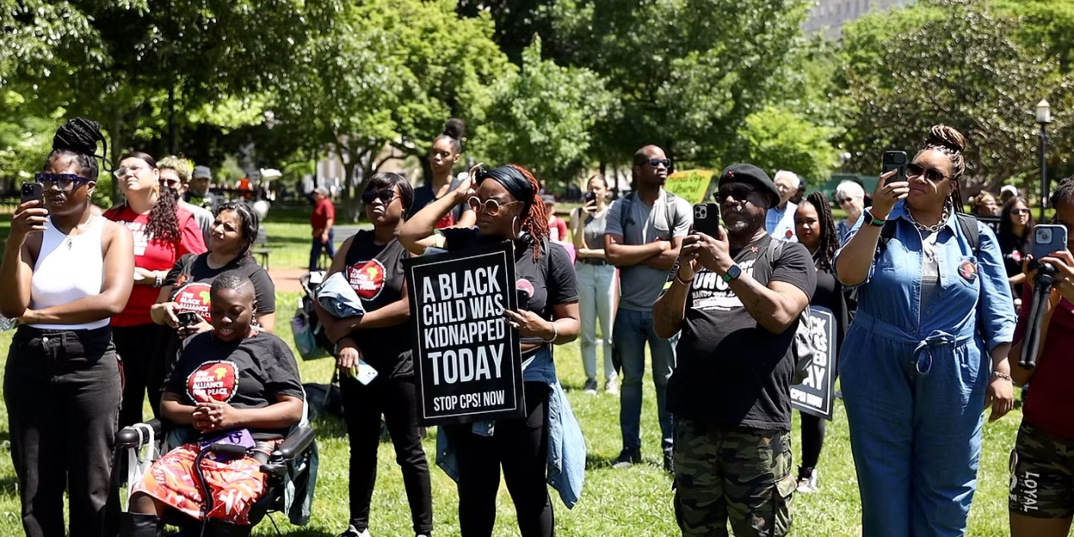 Black parents stand in a park, listening to a speaker. One woman holds a sign that reads "A Black Child Was Kidnapped Today".