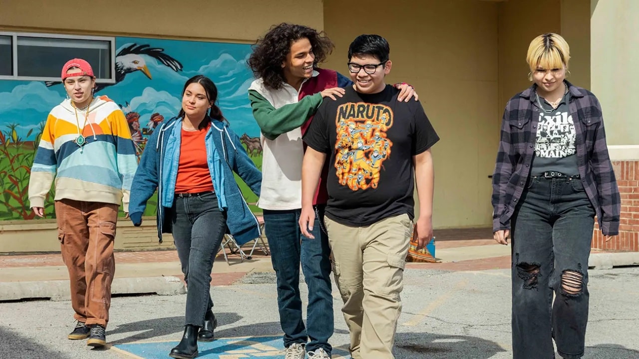 A screenshot of the show “Reservation Dogs” shows five Indigenous teenagers wearing grunge clothes. They are walking out of a community center and smiling. 